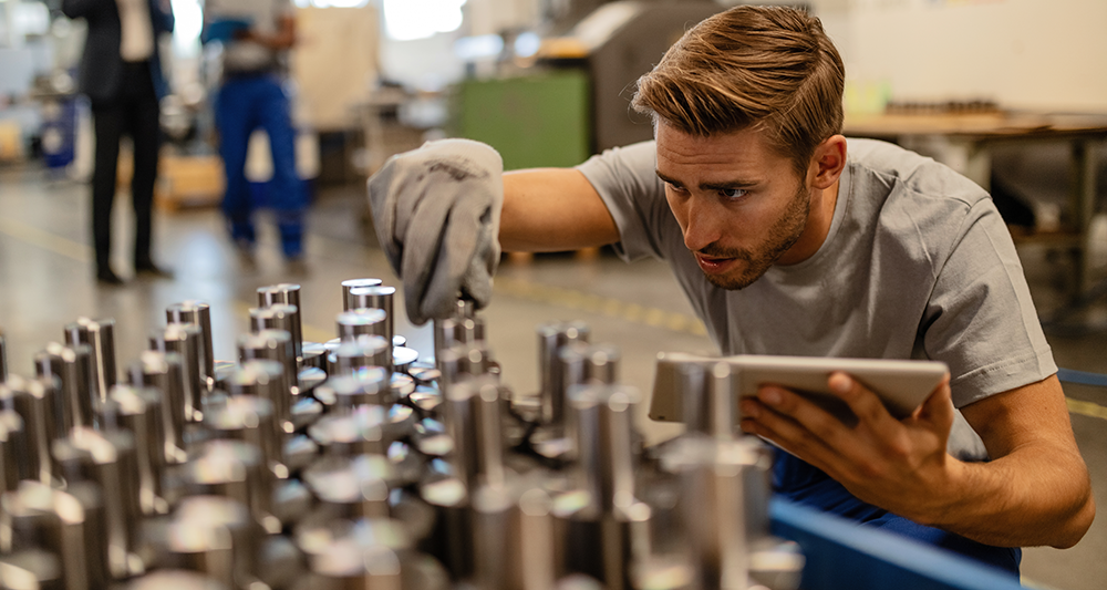 Young steel worker examining manufactured rod cylinder while using digital tablet in distribution warehouse - cropped